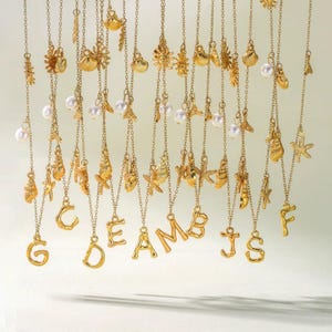 May include: A collection of gold-coloured necklaces featuring seashell, starfish, and pearl pendants. The word "DREAMS" is spelled out in gold letters. Other gold letter charms are also visible.