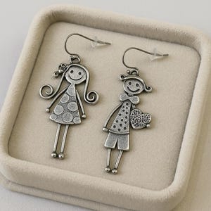 May include: Silver-toned dangle earrings featuring whimsical stick figures of a girl and a boy. The girl has long curly hair and a polka-dot dress. The boy holds a heart-shaped object. Both figures have smiling faces. The earrings are displayed in a beige jewelry box.