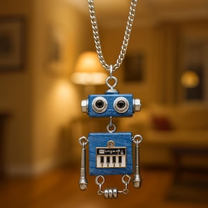 Puede incluir: Colgante de robot azul con una cadena plateada. El robot tiene una cabeza rectangular con dos ojos redondos y un cuerpo rectangular con un diseño de teclado de piano. Los brazos y las piernas son plateados. El colgante cuelga de una cadena plateada.