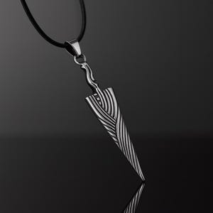 Op de afbeelding: Een zilverkleurige hangerketting met een dolkvormige hanger. De hanger heeft een getextureerd lemmet met een veervormig patroon en hangt aan een zwart koord. De handgreep van de dolk heeft een gebogen ontwerp.