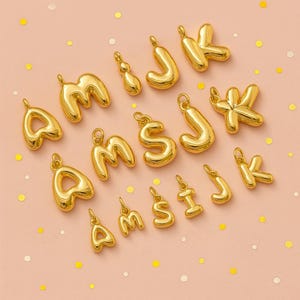 Peut inclure: Charm en forme de lettres de l'alphabet dorées, notamment A, M, S, J et K. Les breloques sont disposées sur une surface rose clair avec des confettis éparpillés.