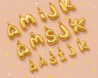 May include: Gold-colored alphabet charms in various letter shapes, including A, M, S, J, and K. The charms are arranged on a light pink surface with scattered confetti.