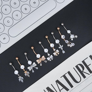 May include: An assortment of belly button rings with various designs. The rings feature silver and gold-toned metal with clear, sparkling gemstones and decorative charms, including bows, leaves, and a planet. The jewelry is displayed on a black surface near a keyboard.