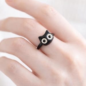 May include: A black cat-shaped ring with white eyes. The ring is a minimalist design, featuring a glossy finish. The cat's head is the focal point, with a simple band. The ring is worn on a finger.