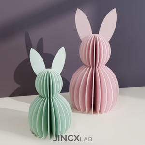 Puede incluir: Dos esculturas decorativas de conejos de papel. Una es verde claro, la otra rosa. Ambas tienen un dise&ntilde;o en capas y acanalado, con orejas largas. Las esculturas est&aacute;n sobre una superficie blanca con un fondo morado y gris. El texto "JINCXLAB" est&aacute; en la parte inferior.