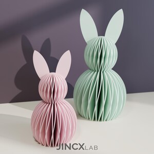 Puede incluir: Dos esculturas decorativas de conejos de papel. Una es rosa, la otra verde menta. Cada conejo tiene un cuerpo esf&eacute;rico en capas y orejas largas y erguidas. Los conejos est&aacute;n sobre una superficie blanca con un fondo morado. El logotipo "JINCXLAB" es visible.