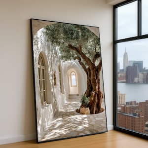 May include: A large rectangular framed wall art print featuring a Mediterranean-style stone corridor with a gnarled olive tree, arched windows, and a stone-paved floor, leaning against a plain wall next to a window overlooking a city skyline.