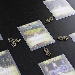 Puede incluir: Una configuración de juego de mesa con varias cartas ilustradas y fichas decorativas de metal. Las cartas tienen ilustraciones y texto, con una carta etiquetada como "Miskatonic Quad". Las fichas son de color dorado y tienen un diseño estilizado.