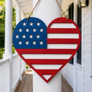 May include: A heart-shaped wooden wall hanging featuring the American flag design. The top left section is blue with white stars, and the right side has red and white stripes. It hangs from a natural-colored rope.