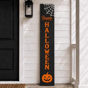 Könnte beinhalten: Ein hohes, rechteckiges schwarzes Holzschild für die Veranda mit dem vertikal angeordneten orangefarbenen Schriftzug "Happy HALLOWEEN". Oben ist ein weißes Spinnennetz abgebildet, unten ein orangefarbenes Kürbis-Gesicht.