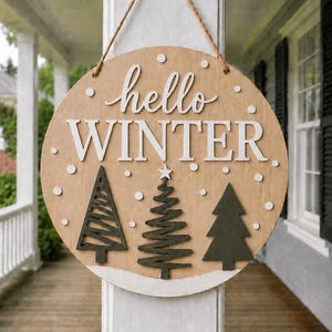 Peut inclure: Un panneau rond en bois avec une finition brun clair portant le texte blanc hello WINTER. Le design comprend trois sapins stylisés vert foncé en bas, des accents circulaires blancs représentant la neige et une corde en jute pour suspendre.