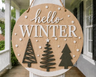 Pode incluir: Uma placa redonda de madeira com acabamento castanho claro apresentando o texto branco hello WINTER. O design inclui três pinheiros estilizados verde-escuros na parte inferior, detalhes circulares brancos representando neve e uma corda de juta.