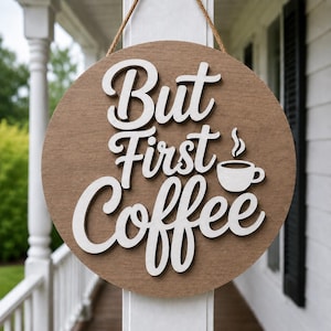 Puede incluir: Cartel de madera redondo con la frase "But First Coffee" en letras blancas estilizadas. Incluye un pequeño gráfico de taza de café. El cartel es marrón con un aspecto de veta de madera natural y está colgado con una cuerda marrón.