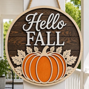 May include: A circular wooden wall sign featuring a dark brown horizontal plank background. The text "Hello FALL" is displayed in white 3D lettering above a large orange pumpkin cutout surrounded by light-colored wooden leaf accents.