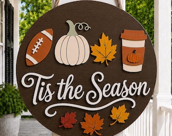 May include: A round dark brown wooden hanging sign with a rope hanger. It features a football, a white pumpkin, a coffee cup with a pumpkin icon, and autumn leaves. The text "Tis the Season" is written in white script lettering across the center.