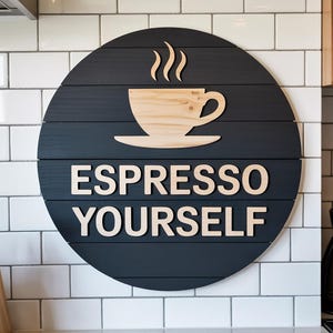 May include: A circular, black wooden sign featuring the phrase "ESPRESSO YOURSELF" in light-coloured lettering. A light-coloured wooden coffee cup and steam design is at the top. The sign is mounted on a white tiled wall.