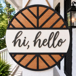 May include: A circular wooden door sign featuring a natural wood grain finish with black geometric sunburst patterns at the top and bottom. A central white horizontal band displays the black script text "hi, hello" and a rope hanger is attached.