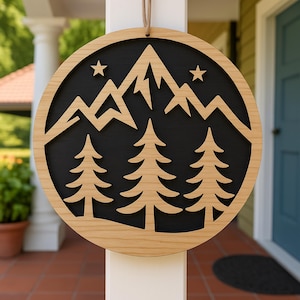 May include: A circular wooden sign featuring a mountain and tree design. The sign has a black background with a mountain range and two stars cut out. Two pine trees are at the bottom. It is suspended by a natural fibre cord.