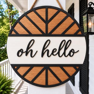 May include: A circular wooden door sign featuring a black frame with geometric sunburst patterns in natural wood grain. A horizontal white centre panel displays the black 3D script text "oh hello". The sign hangs from a thick tan rope.