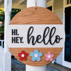 May include: A circular wooden sign with a light brown top and bottom, and a white central section. The sign features the words "Hi. Hey. hello" in black, with three colourful flower accents. A decorative item for the home.