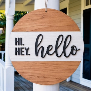 May include: A circular wooden sign with a natural wood grain top and bottom, and a white central band. The sign displays the words "HI. HEY." in black block letters and "hello" in a black cursive font. A jute string is attached for hanging.