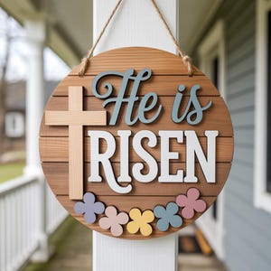 May include: A round wooden sign with the words "He is RISEN" in white and blue lettering. A light-colored cross is on the left, and colorful flower cutouts are along the bottom. The sign hangs from a jute rope.