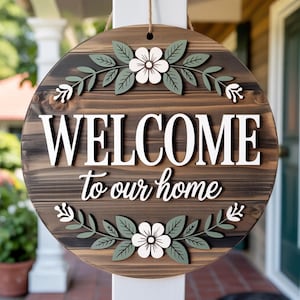 May include: A round wooden sign with a dark brown stained finish featuring the white text WELCOME to our home. The sign is decorated with two white flowers and sage green leaves at the top and bottom, and it hangs from a thin brown twine cord.