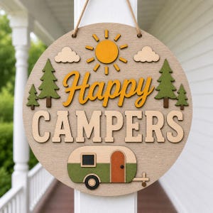 May include: Round wooden sign with the words "Happy Campers" in orange and natural wood tones. The sign features a sun, clouds, trees, and a camper trailer. It hangs from a jute rope.