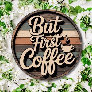 Könnte beinhalten: Rundes Holzschild mit dem Schriftzug "But First Coffee" in einer stilisierten Schriftart. Das Schild zeigt eine Kaffeetassen-Grafik und einen Hintergrund aus horizontalen Holzmaserungsstreifen in Braun-, Grau- und Beigetönen. Das Schild ist von weißen Blumen und grünen Blättern umgeben.