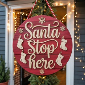 May include: A round, red wooden sign with the words "Santa Stop Here" in cream-colored, raised lettering. The sign features snowflakes and stockings. A red and white striped rope is used for hanging.