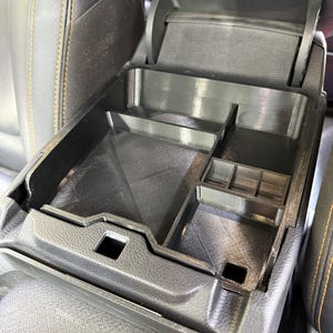 May include: Black plastic car console organiser with several compartments. The organiser is designed to fit inside a car's centre console, providing storage for small items. The car's interior has black leather seats with yellow stitching.