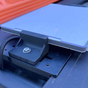 May include: A black 3D-printed device with a silver tablet secured on top. The device is mounted on a black track system. The text "Dane 3D Labs" is visible on the side of the device. A black cable is connected to the device.