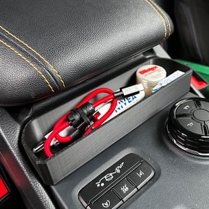 May include: A black car console organizer with a red USB cable, a tube of Nivea cream, and a small container of Blistex lip balm. The organizer sits next to a black control dial and a black leather armrest with yellow stitching.