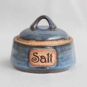 May include: A blue ceramic salt container with a lid and a handle. The container has a brown label that says "Salt".