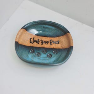 May include: A round, ceramic soap dish featuring a speckled teal and tan glaze. The center of the dish displays the black text Wash Your Paws, accompanied by several small, embossed paw print designs arranged in a semi-circle below the lettering.