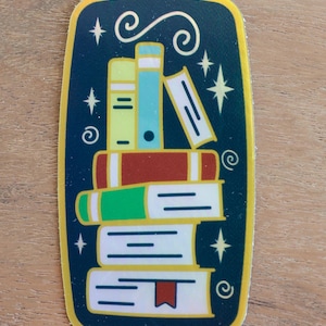 May include: A sticker featuring a stack of books in various colors, including green, red, and blue, against a dark blue background. The design includes stars and swirls, with the text "Nazzeramics Pottery" at the bottom.
