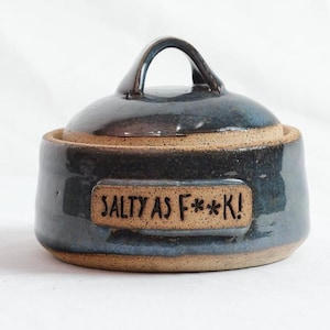 May include: A round, dark blue glazed ceramic salt cellar with a matching lid and a handle. The front features a rectangular, unglazed tan patch with the text SALTY AS F**K! stamped in black lettering. The base is also unglazed tan ceramic.