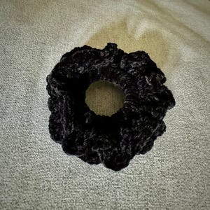 May include: A black scrunchie made of a soft, textured material. The hair accessory is circular with a hole in the center. The scrunchie is a solid black color and appears to be made of a plush fabric.