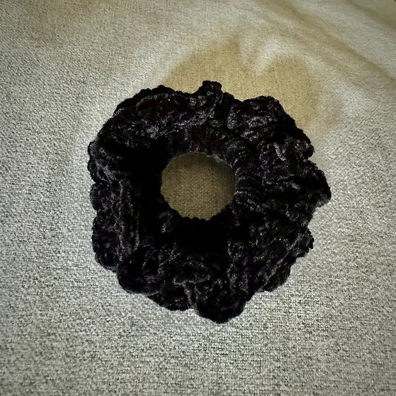 May include: A black scrunchie made of a soft, textured material. The hair accessory is circular with a hole in the center. The scrunchie is a solid black color and appears to be made of a plush fabric.