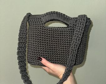 May include: A dark gray crocheted handbag with a handle and a long strap. The bag has a rectangular shape and a textured appearance. The strap is a twisted rope design. The bag is held up by a hand.