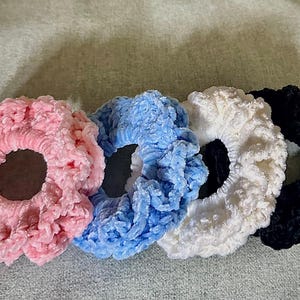 May include: Four plush scrunchies in a row. The scrunchies are in shades of pink, blue, white, and black. The fabric appears to be a soft, velvety material. These hair accessories are a simple way to add color to any outfit.