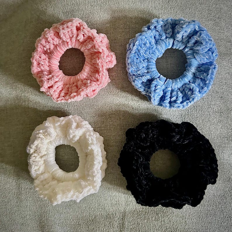 May include: Four plush scrunchies in pastel colors. The scrunchies are in pink, blue, white, and black. The scrunchies are made of a soft, velvety material. These hair accessories are perfect for adding a touch of color to any hairstyle.