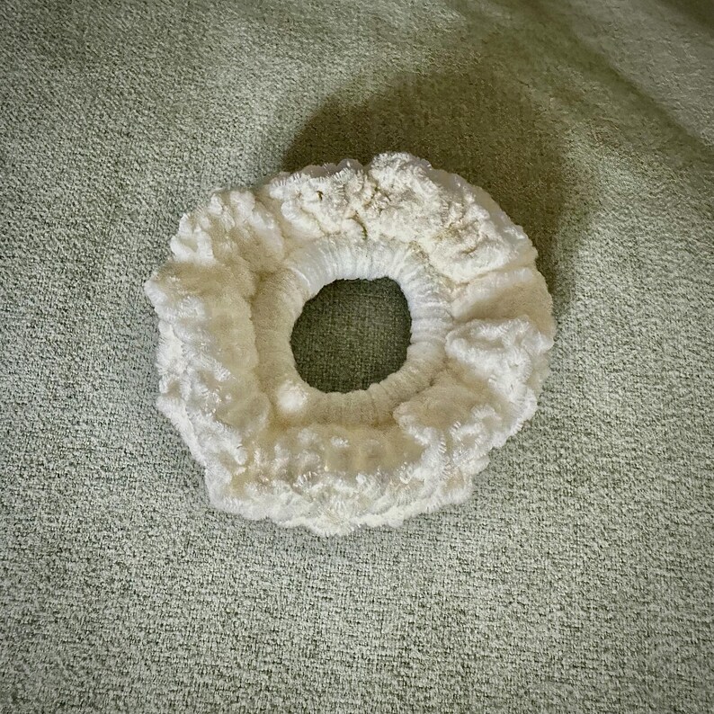 May include: A fluffy, off-white scrunchie with a large center opening. The scrunchie is made of a soft, textured material, and is circular in shape. The background is a light green, textured surface.
