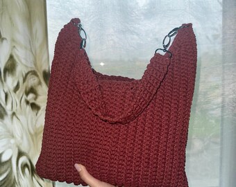 May include: A burgundy crochet tote bag with a chain strap. The bag is made of a thick, textured yarn and has a rounded top. The chain strap is silver-colored. The bag is held up by a hand.