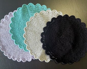 May include: A collection of five crocheted placemats in a variety of colors. The placemats are round with a scalloped edge. Colors include lavender, teal, cream, and black. The placemats are handmade and have a textured appearance.