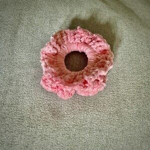 May include: A light pink scrunchie made of a soft, textured material. The scrunchie is circular with a hole in the center and a ruffled edge. The background is a light green fabric.