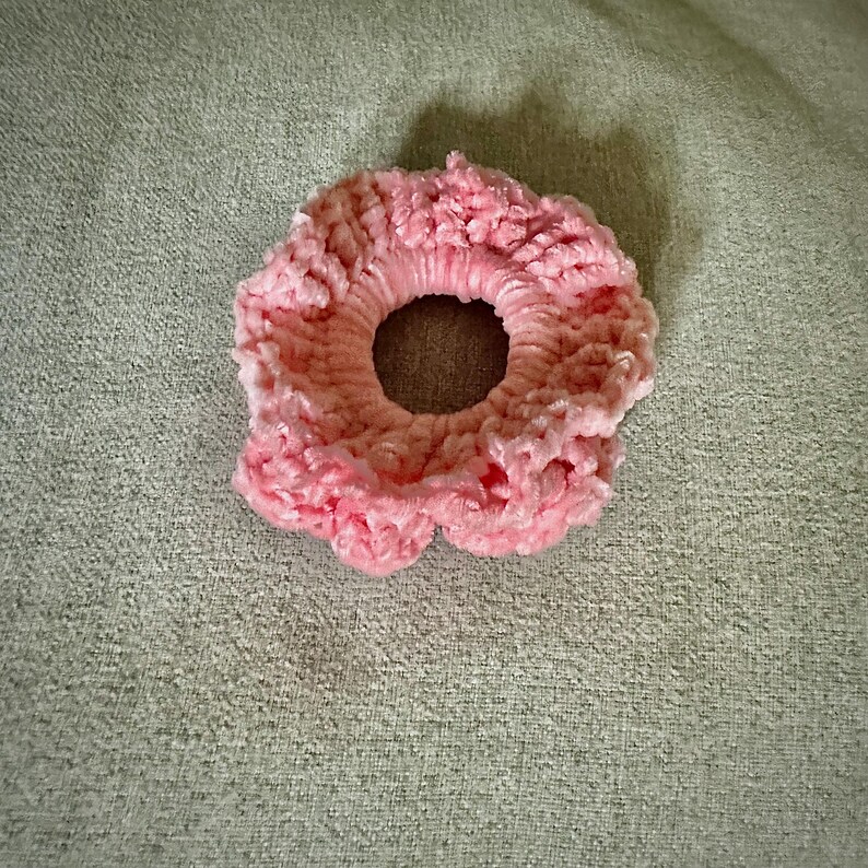 May include: A light pink scrunchie made of a soft, textured material. The scrunchie is circular with a hole in the center and a ruffled edge. The background is a light green fabric.