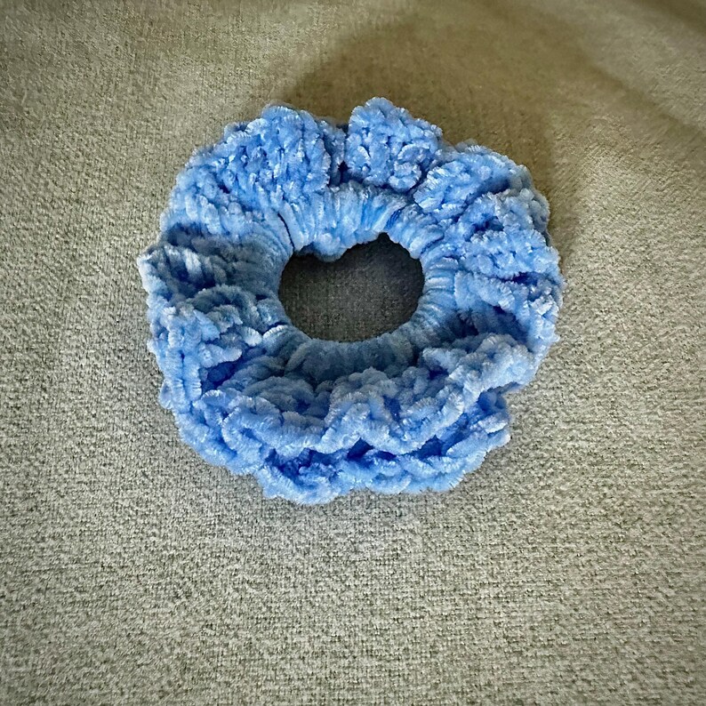 May include: A light blue scrunchie made of a soft, plush material. The hair accessory is circular with a hole in the center. The scrunchie is a solid color and appears to be made of a velvet-like fabric.