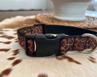 May include: A dog collar with a black plastic buckle and a D-ring. The collar features a brown and black leopard print pattern on a black nylon base. The collar is designed for pets.