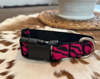 Könnte beinhalten: Schwarzes Hundehalsband mit einem pinkfarbenen Zebra-Streifenmuster. Der Kragen hat eine schwarze Kunststoffschnalle und einen silbernen Metall-D-Ring. Das Halsband liegt auf einer braun-weiß gefleckten Oberfläche.